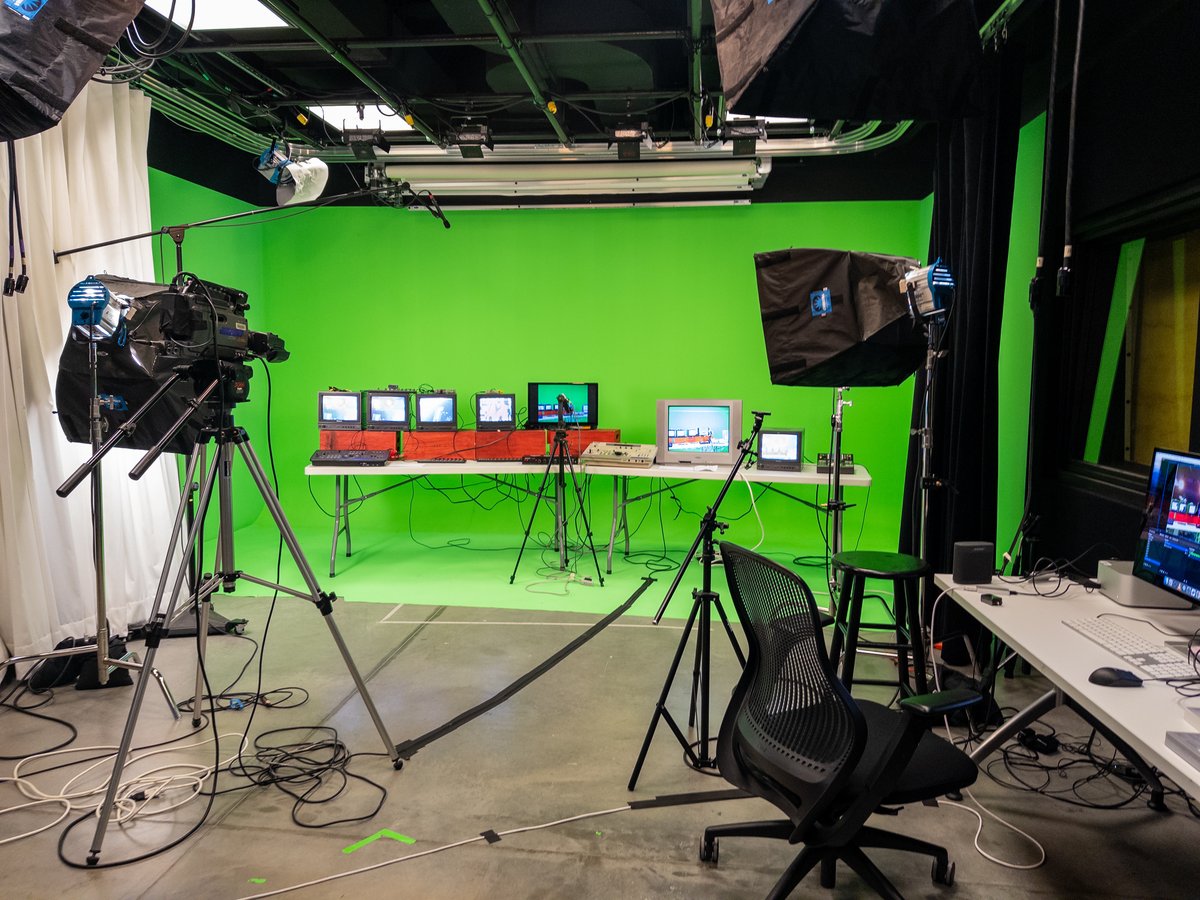 a greenscreen studio with a bunch of old TVs and analog video equipment set up in front of some lights and a streaming computer