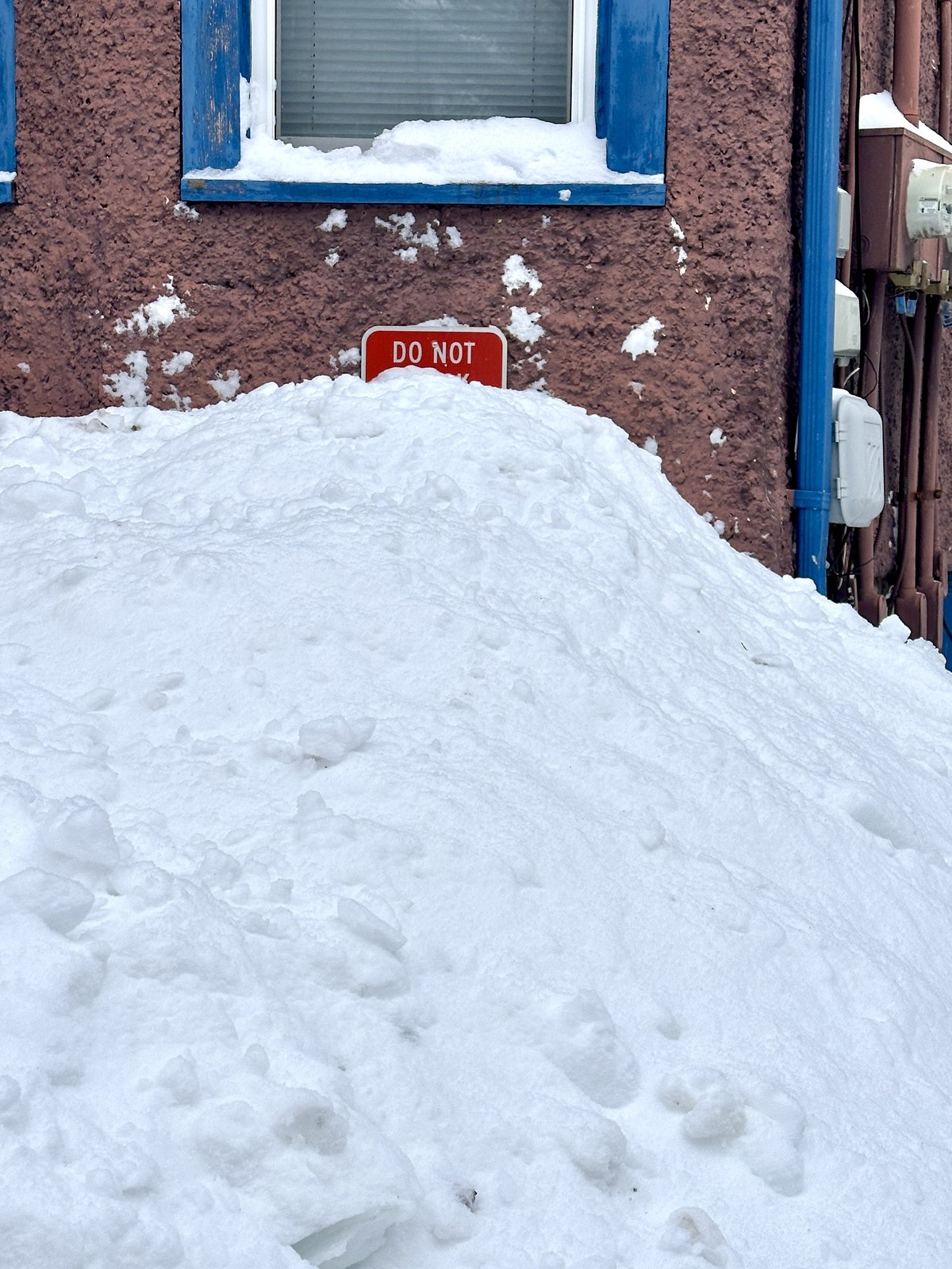 snow piled up against the side of a building with purple stucco walls. the sign says "do not" and the rest is obscured by the snow