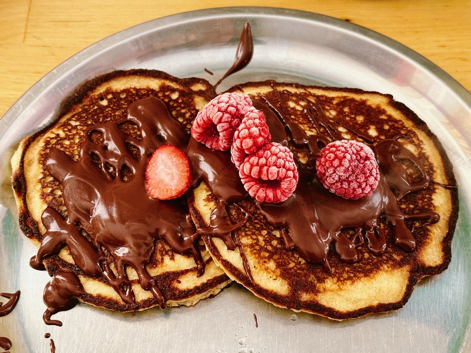 two beautiful pancakes on a silver plate, covered in thick chocolate syrup, and topped with frozen raspberries