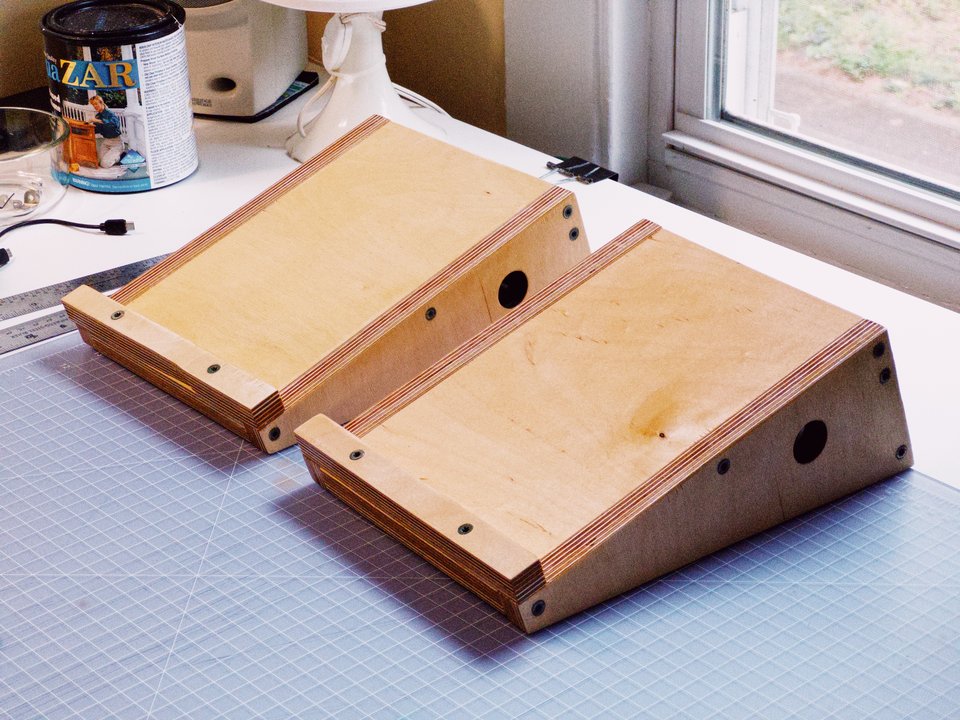 two wedge-shaped speaker stands made from thick plywood, sitting on a table next to a window