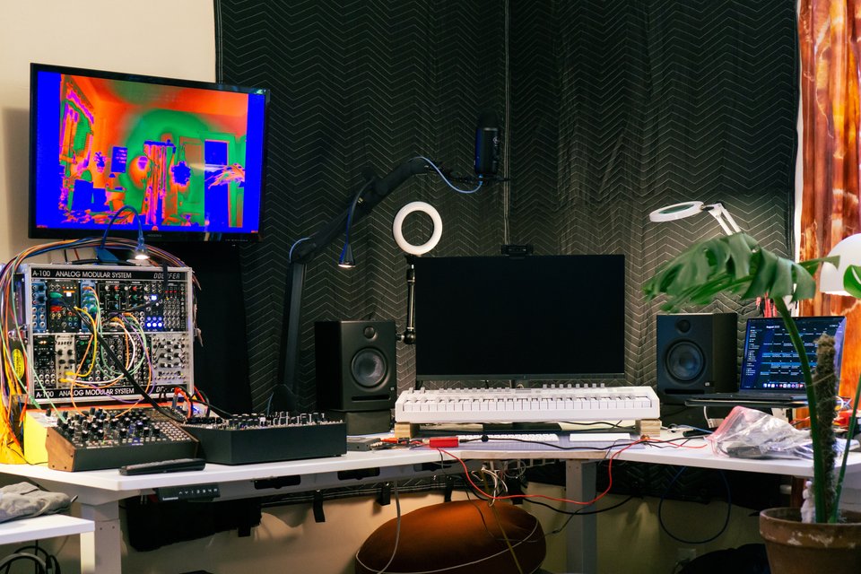 a corner desk with a computer, external monitor, midi keyboard, monitor speakers, a modular synthesizer, and a tv screen showing a colorized video feed