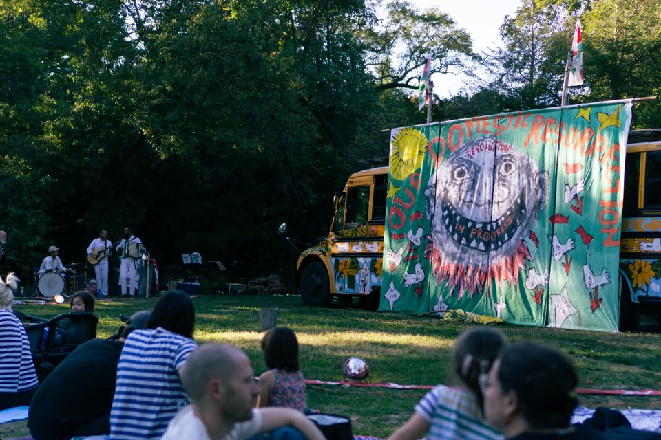 the set of the bread and puppet circus, a yellow school bus painted with murals and some people playing accordion and tuba