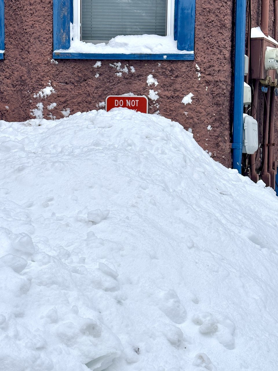 snow piled up against the side of a building with purple stucco walls. the sign says "do not" and the rest is obscured by the snow